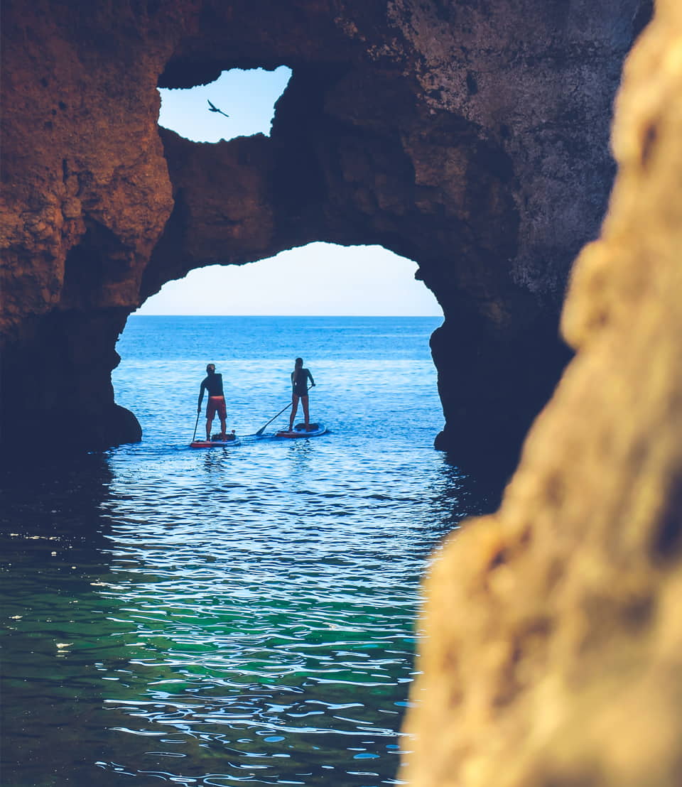 A couple paddle surfing past a rock arch
